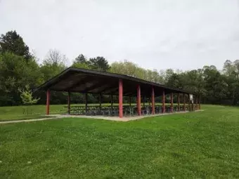 Open-sided picnic shelter with red posts and rows of picnic tables beside a grassy field and trees under an overcast sky.