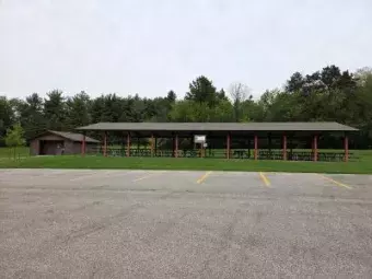 Large covered picnic pavilion with red support posts, picnic tables, and a nearby parking lot surrounded by trees.