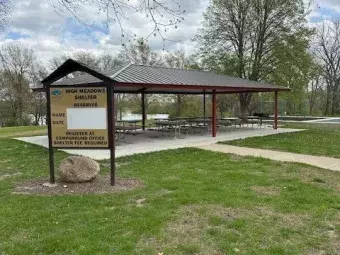 This photo depicts cement walkway leading up to a steel picnic shelter structure with multiple picnic tables and a grill.