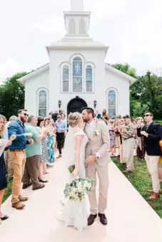 Front view of the church with wedding guests blowing bubbles towards a kissing bride and groom
