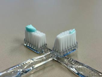Two toothbrushes - one shows a pea-sized amount of toothpaste, the other shows a rice-grain sized amount of toothpaste.