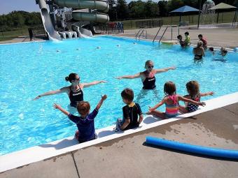 Two instructors in the pool demonstrating a skill to a group of children sitting along the edge of the pool.