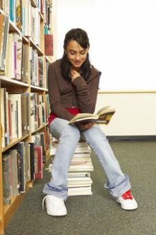 Girl reading while sitting atop stack of books in library