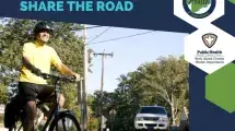Bike and pedestrian safety awareness banner with text “Bike & Ped Safety: Share the Road,” featuring a smiling adult cyclist wearing a helmet and yellow shirt riding a bicycle on a suburban street, with a car approaching in the background, trees and utility poles visible, and official logos for Scott County Health Department and Rock Island County Public Health Department on a blue and green graphic layout.