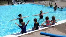 Two instructors demonstrating a skill to children sitting on the side of the pool.